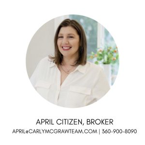 Headshot of April Citizen, Broker, in a circular frame with text below reading "April Citizen, Broker" and contact info "april@carlymcgrawteam.com | 360-900-8090". She is a smiling woman with shoulder-length brown hair, wearing a white button-down shirt with chest pockets and layered gold necklaces, set against a bright indoor background with a blurred vase of flowers.
