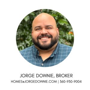 Headshot of Jorge Downie, Broker, in a circular frame with text below reading "Jorge Downie, Broker" and email "homes@jorgedownie.com". He is a smiling, bald man with a dark beard and mustache, wearing a blue plaid button-down shirt against a blurred background of green foliage.
